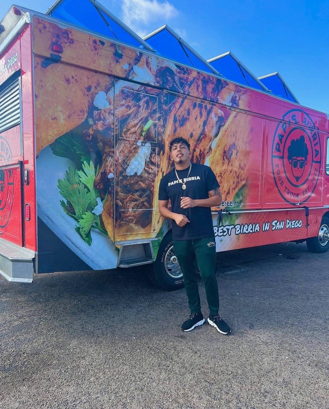 Ivan Cruz, founder and owner of Papi's Birria, standing in front of the food truck, whose side is wrapped in a photo of birria tacos with the badge and Best Birria in San Diego lettering.